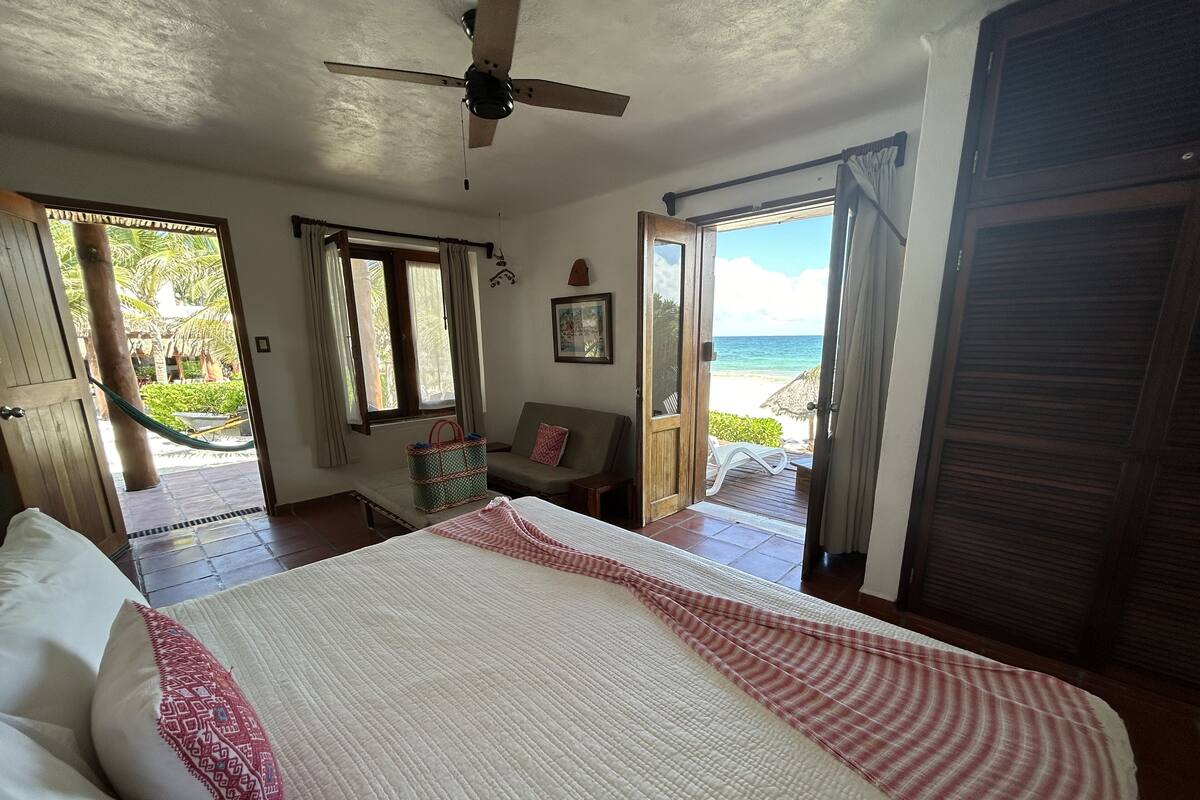 A bedroom with a bed, a ceiling fan, a window with curtains, a door leading to a balcony, and a view of the beach.