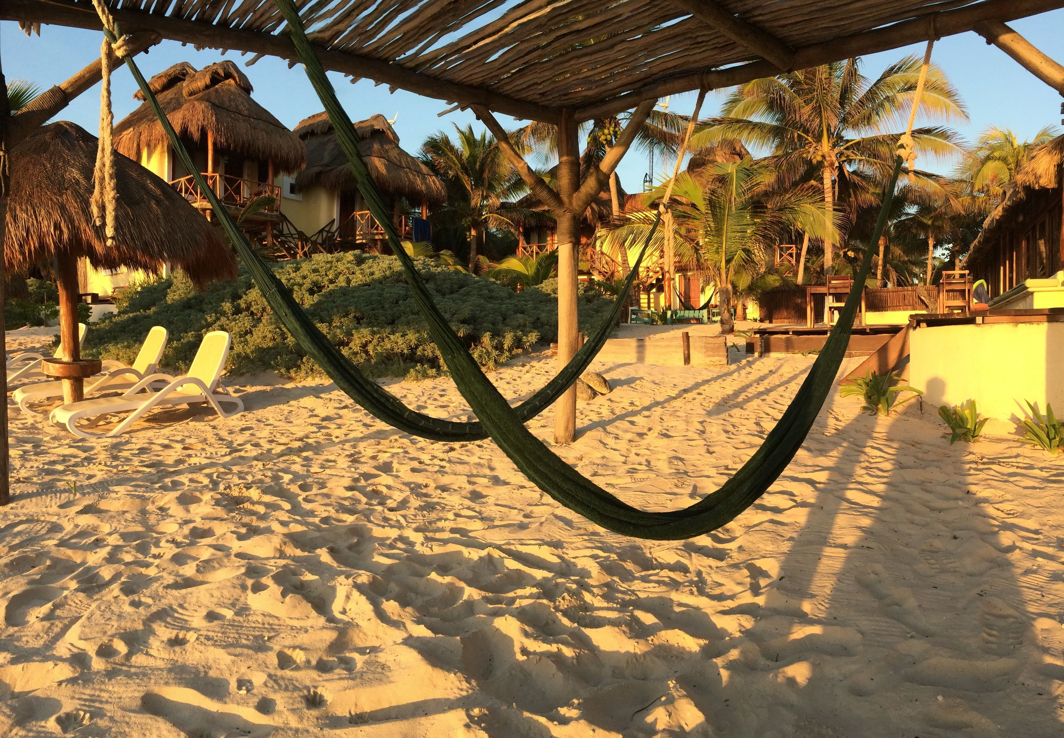 A hammock under a wooden structure on a sandy beach with palm trees and thatched-roof buildings in the background.