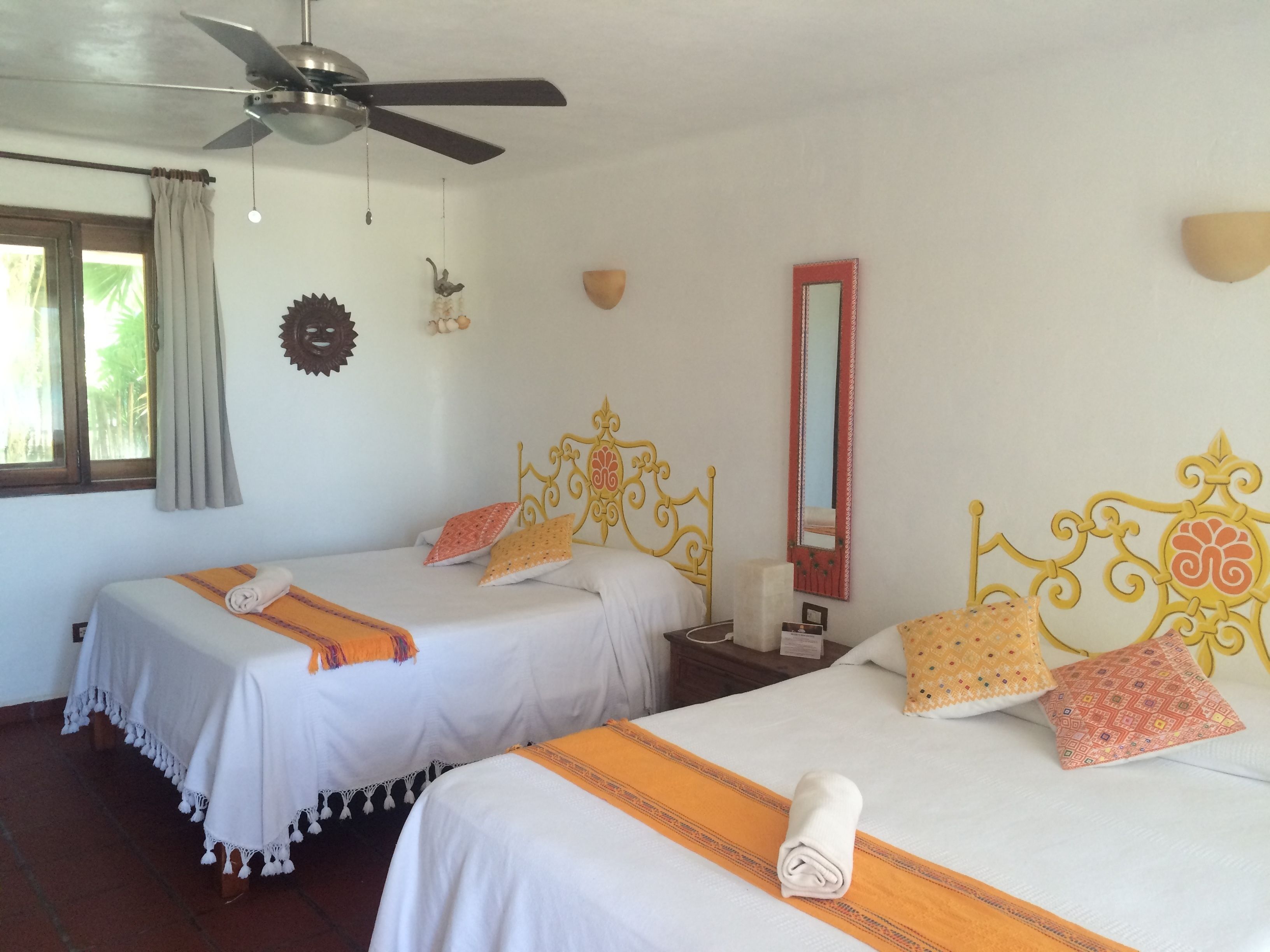 A hotel room with two beds, a ceiling fan, a window with curtains, a wall-mounted mirror, and a decorative wall piece.