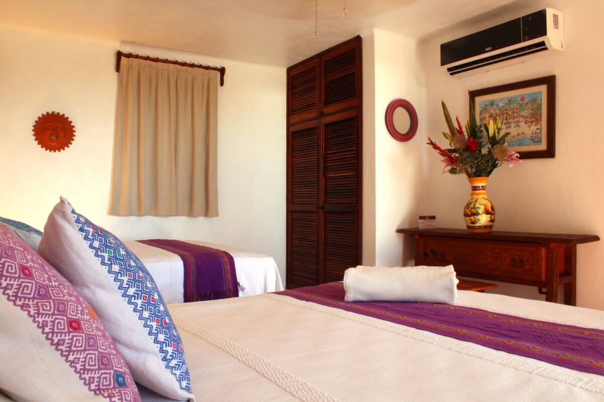 A hotel room with a bed, a wooden wardrobe, a desk with a vase of flowers, and a painting on the wall.