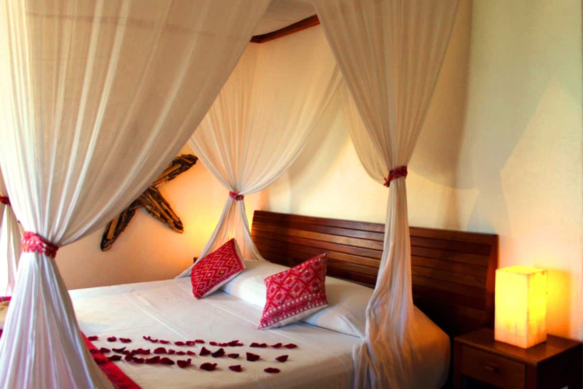 A bed with a canopy, red pillows, and rose petals on the bed.