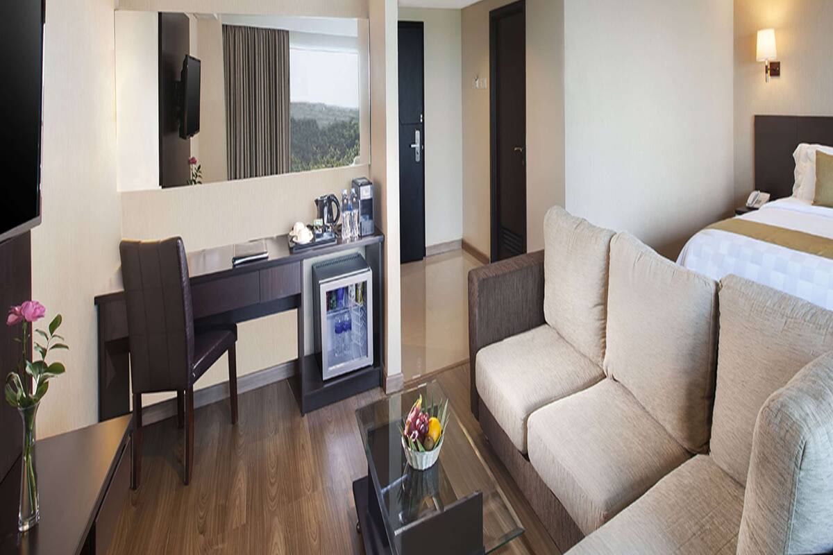 A hotel room with a bed, a sofa, a desk with a computer, a coffee machine, and a TV.