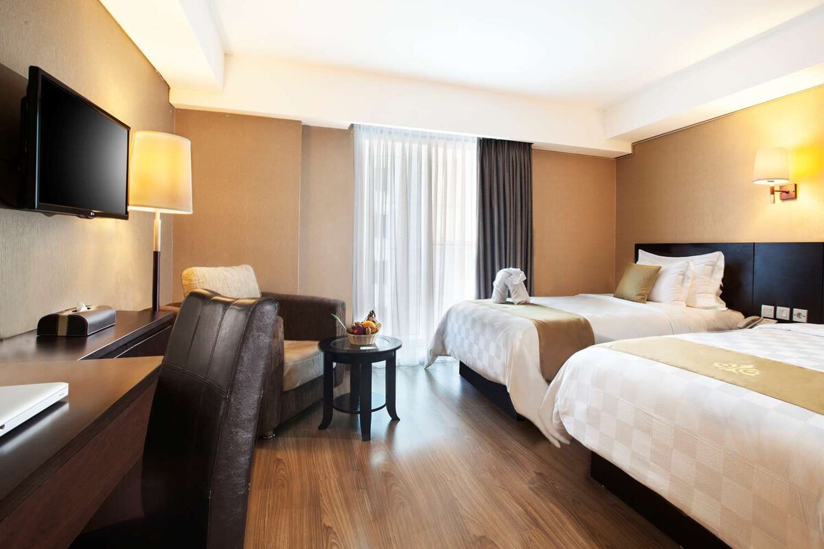 A hotel room with a desk, two beds, a chair, a TV, and a window with curtains.