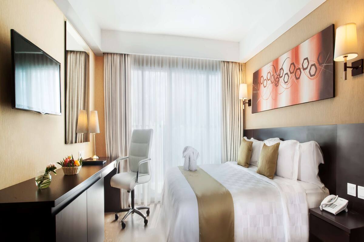 A hotel room with a bed, desk, chair, and a painting on the wall.