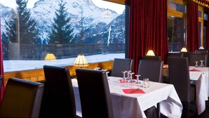 Un restaurante con mesas dispuestas para comer, amplias ventanas con vistas a montañas nevadas y cortinas rojas.