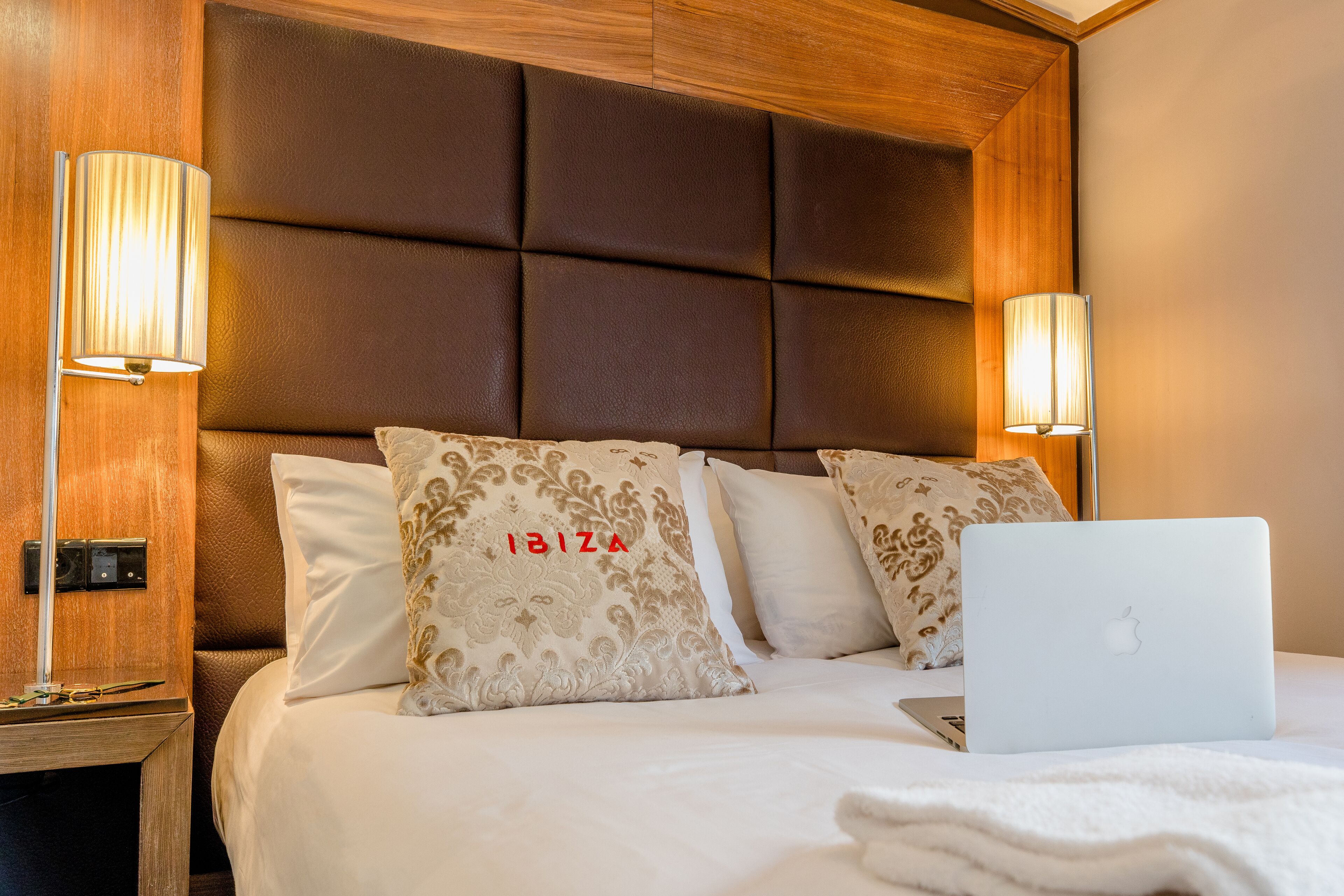 Un lit bien fait, avec un ordinateur portable, deux coussins décoratifs et une tête de lit en bois.