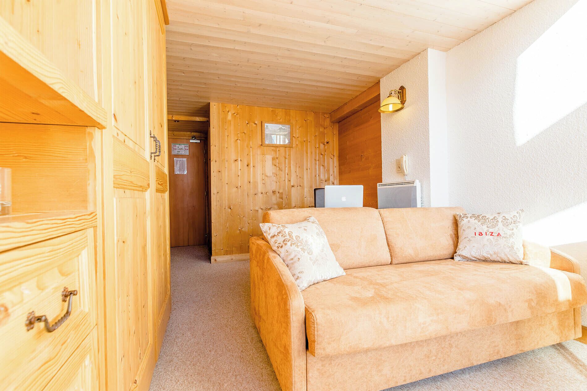 A cozy room with a wooden interior, a sofa with two pillows, a wall-mounted lamp, and a door leading to another room.