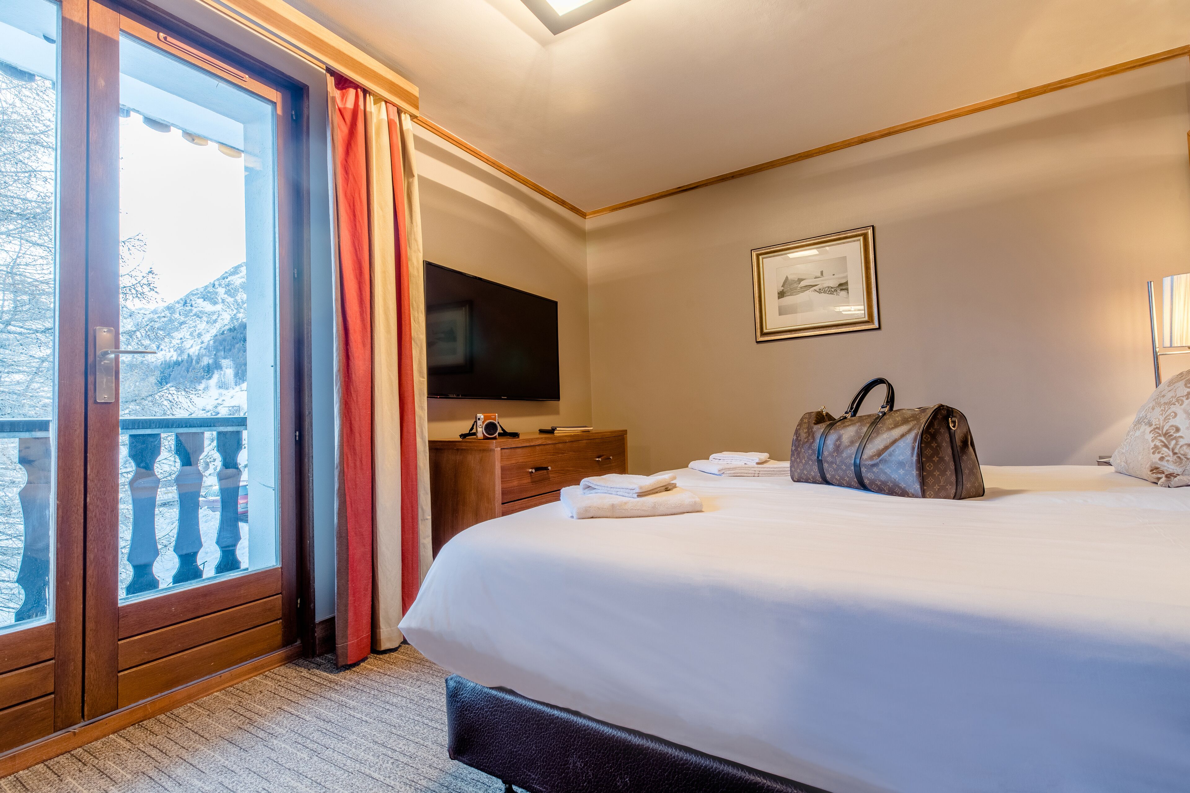 A hotel room with a large bed, a flat-screen TV, a wooden dresser, and a balcony with a view of snow-covered mountains.