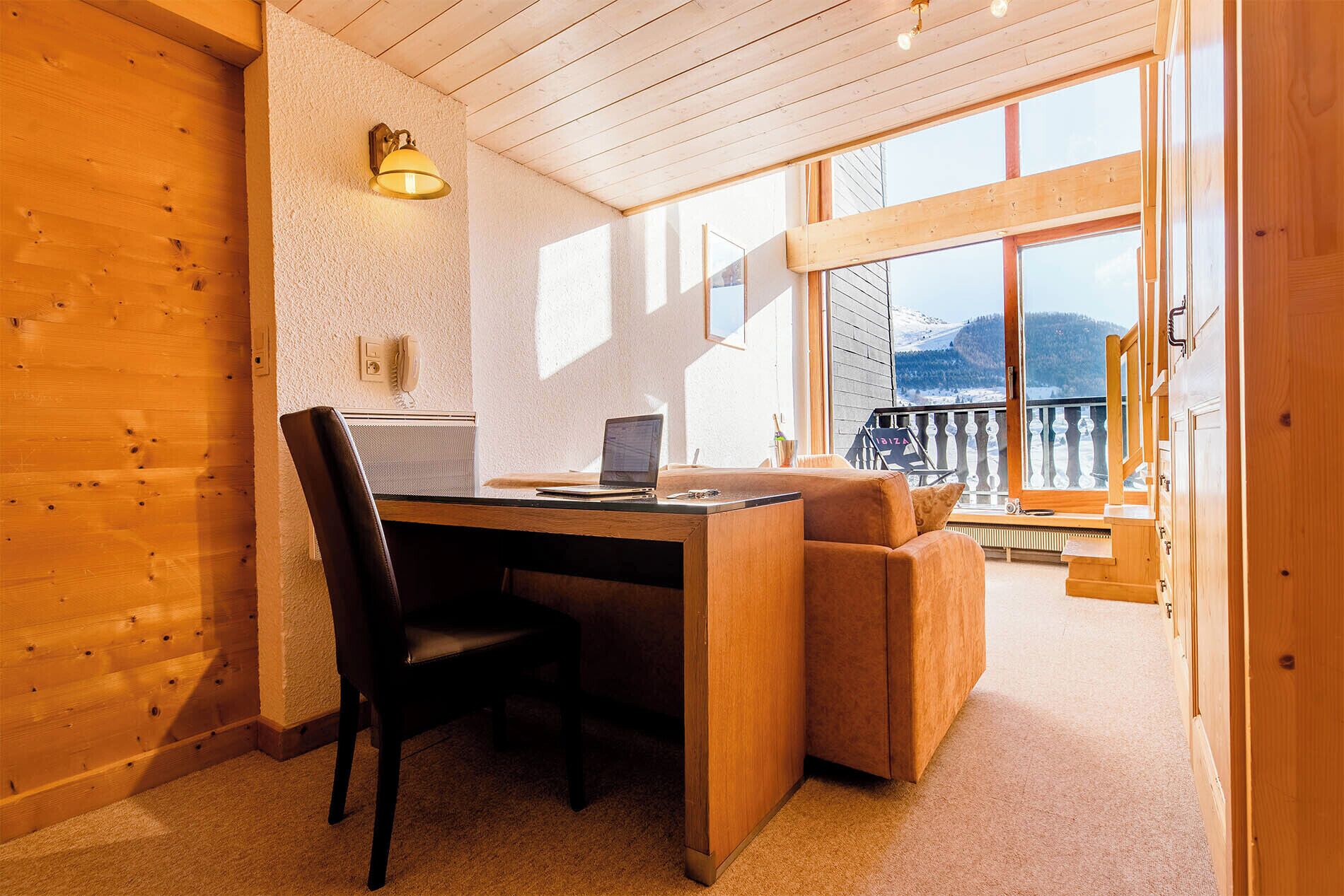 A room with a wooden desk, a chair, a laptop, and a view of a snowy landscape through large windows.