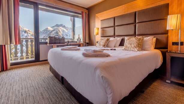 Una habitación de hotel con una cama grande, vista a las montañas, cabecera de madera y una mesita de noche con lámpara.