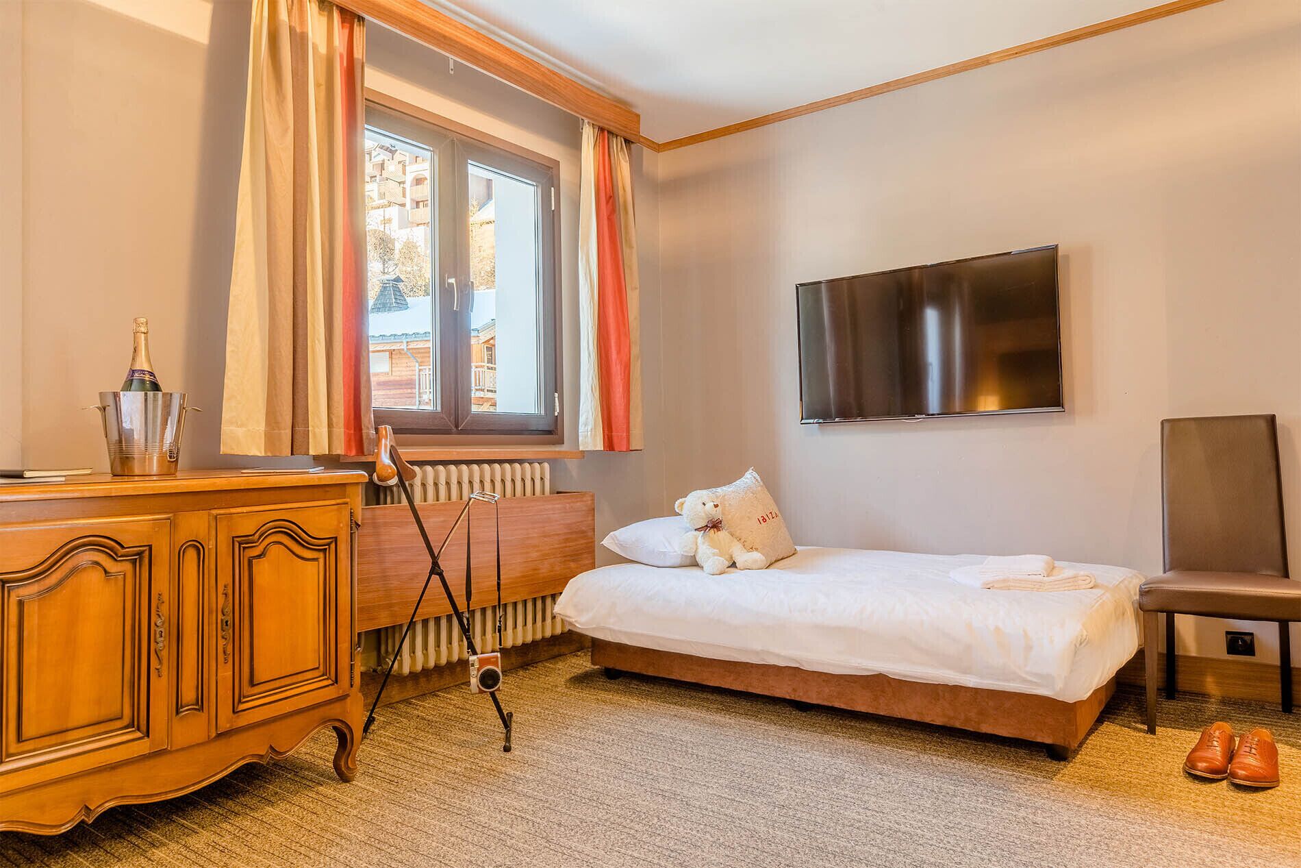 Une chambre d’hôtel avec un lit, une commode en bois, une chaise, un téléviseur et une fenêtre avec des rideaux.