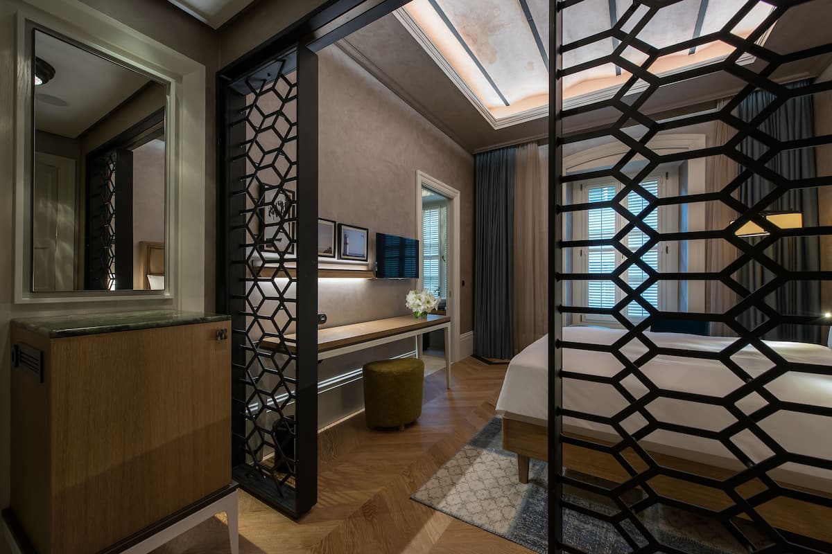 Une chambre d’hôtel moderne avec un meuble en bois, un bureau agrémenté d’un vase de fleurs, un lit doté d’un paravent aux motifs géométriques et un grand miroir.