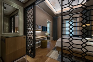 A modern hotel room with a wooden cabinet, a desk with a vase of flowers, a bed with a geometric-patterned screen, and a large mirror.