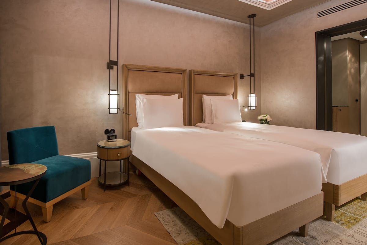 Une chambre d’hôtel avec deux lits, un fauteuil bleu, une petite table ronde et une table de chevet avec une radio.