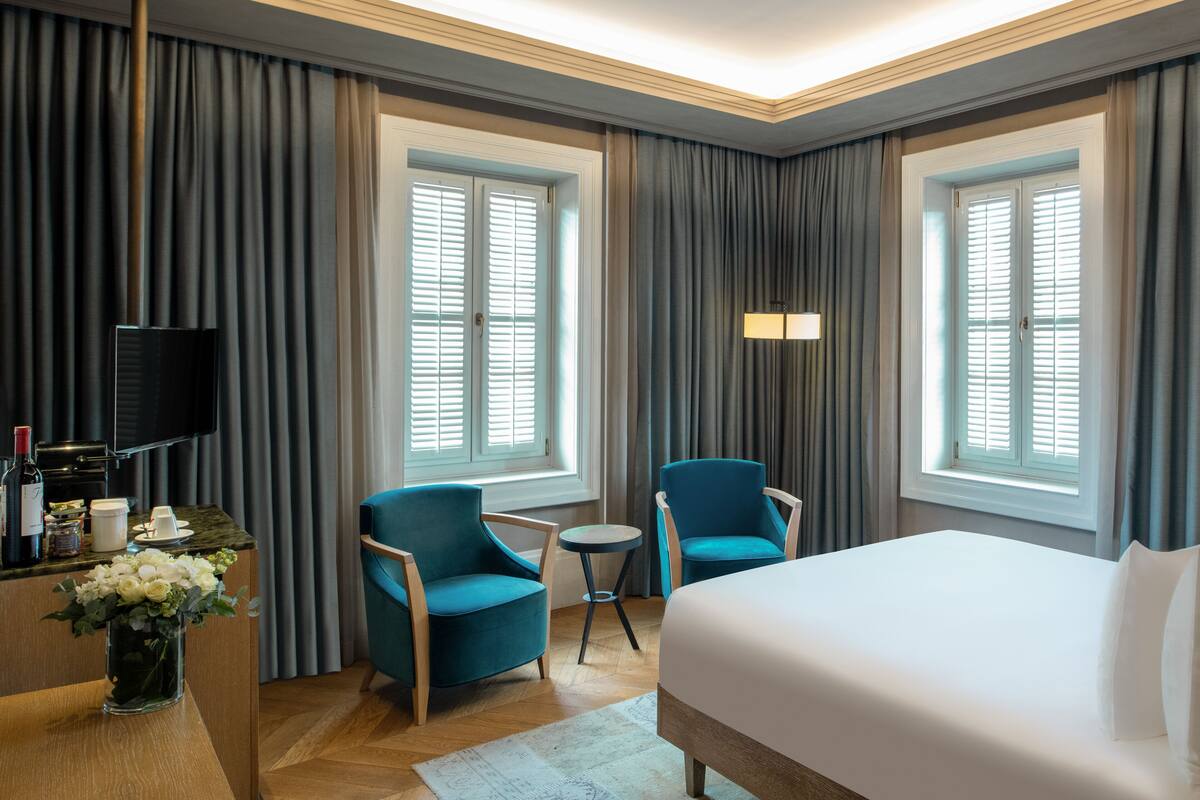 Une chambre d’hôtel avec un grand lit, deux fauteuils bleus, une petite table ronde, un téléviseur et un vase de fleurs.