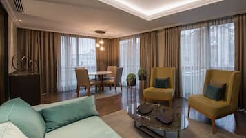 Una habitación de hotel con cama, sofá, mesa de centro, mesa de comedor con sillas y un gran ventanal con cortinas.