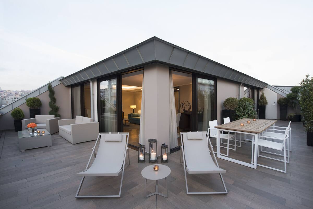 Une terrasse sur le toit au design moderne, comprenant un espace repas avec une table et des chaises, des fauteuils de relaxation et un espace clos par des baies vitrées offrant une vue sur la ville.