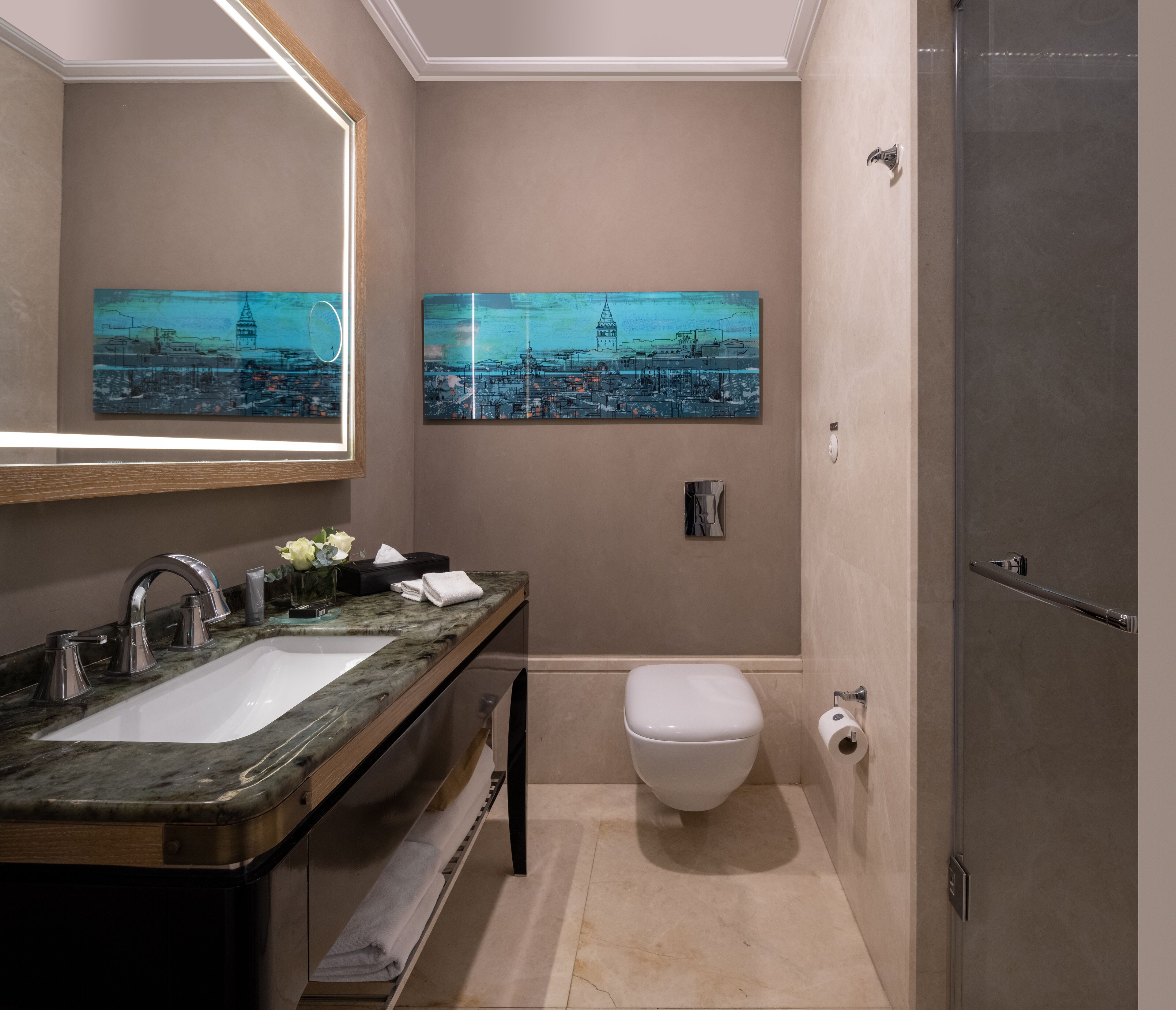 A modern bathroom with a dual sink vanity, a toilet, and a shower. There are two framed cityscape paintings on the wall.