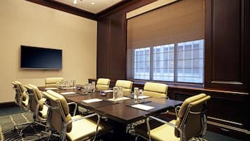 A conference room with a large table, multiple chairs, a flat-screen TV, and a large window with blinds.
