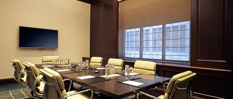 A conference room with a large table, multiple chairs, a flat-screen TV, and a large window with blinds.
