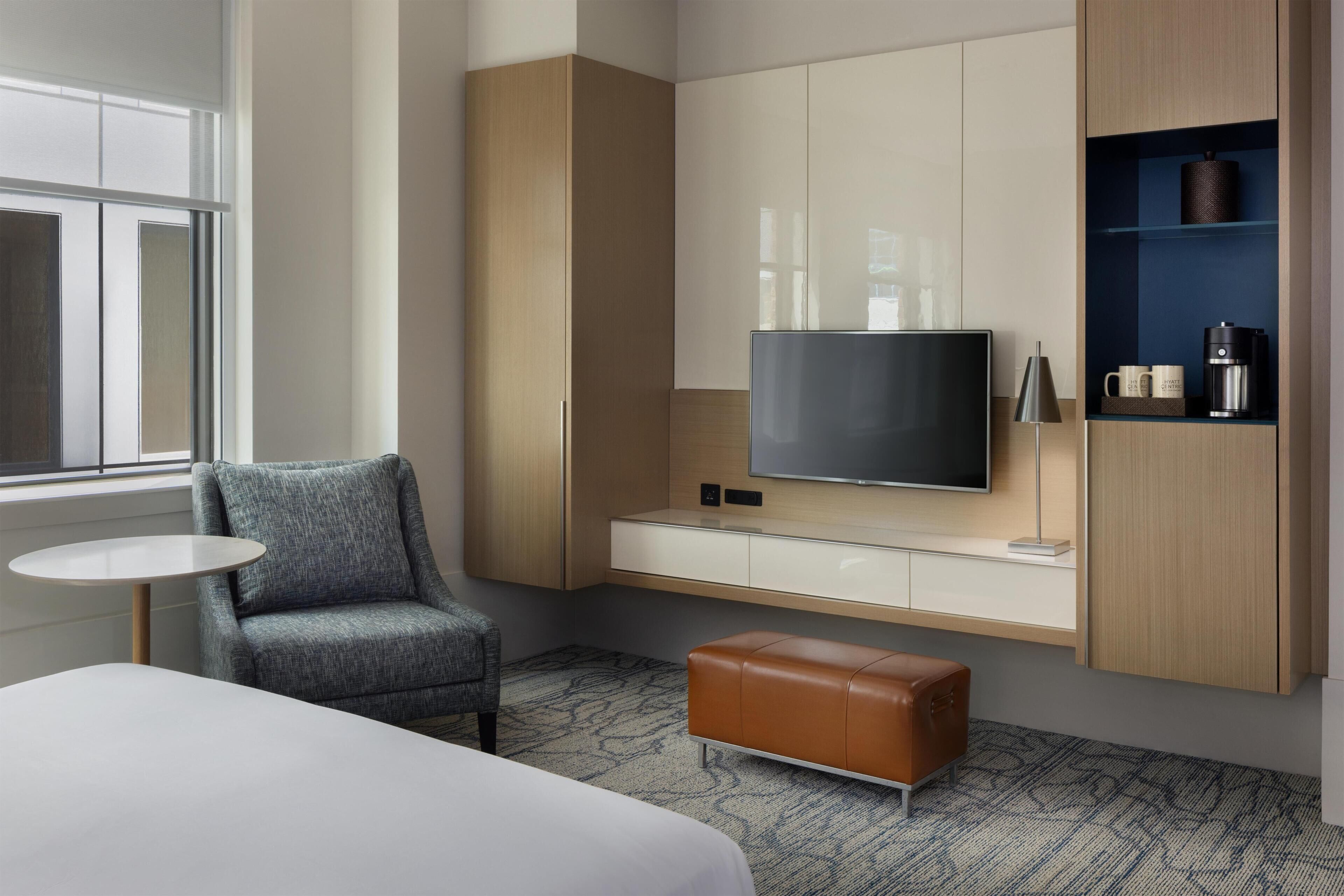 Habitación de hotel moderna con televisor de pantalla plana, zona de estar con sillón y reposapiés, y un mini bar.