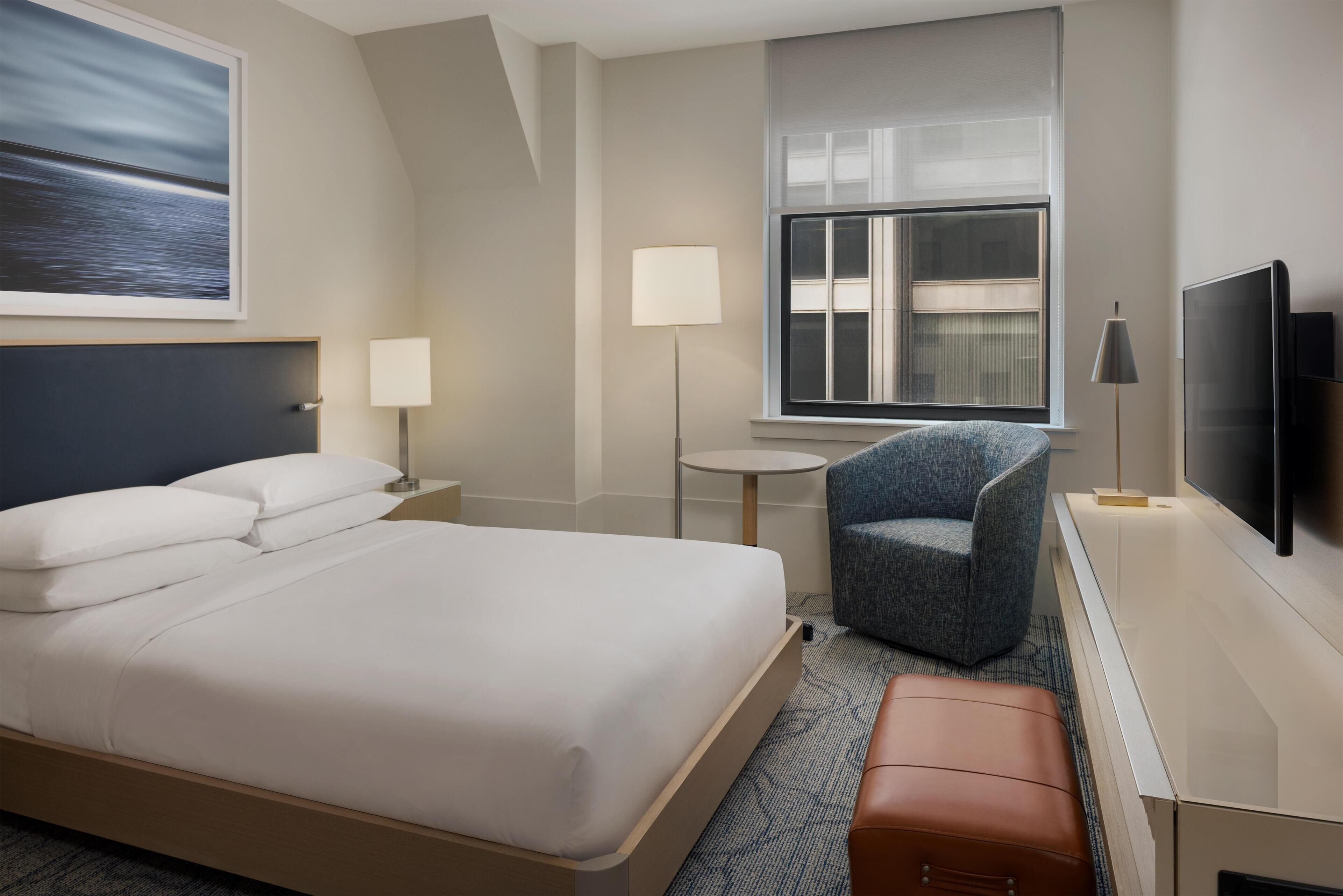 A hotel room with a bed, a chair, a desk with a TV, a window, and a lamp.