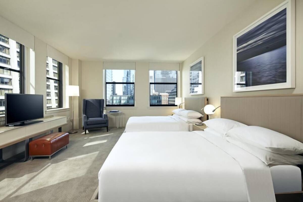 A hotel room with two beds, a TV, a desk, a chair, and large windows with a city view.