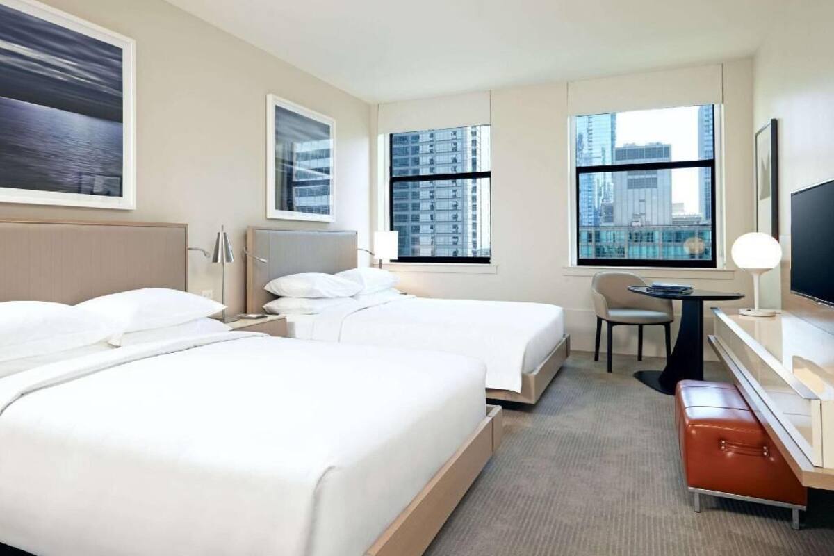 A hotel room with two beds, a desk, a chair, a TV, and large windows with a city view.