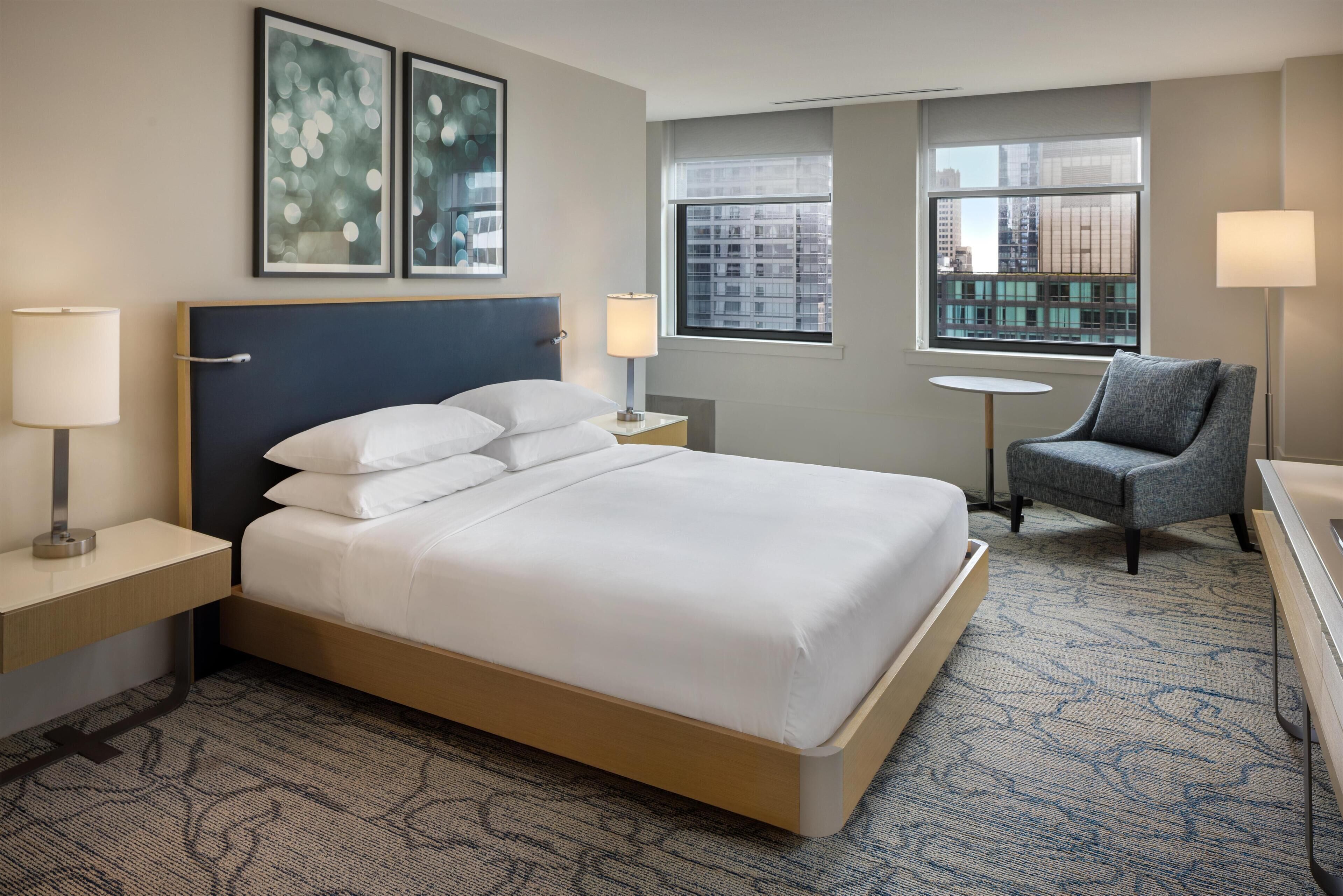 A hotel room with a bed, two bedside lamps, a chair, a small table, and artwork on the wall.