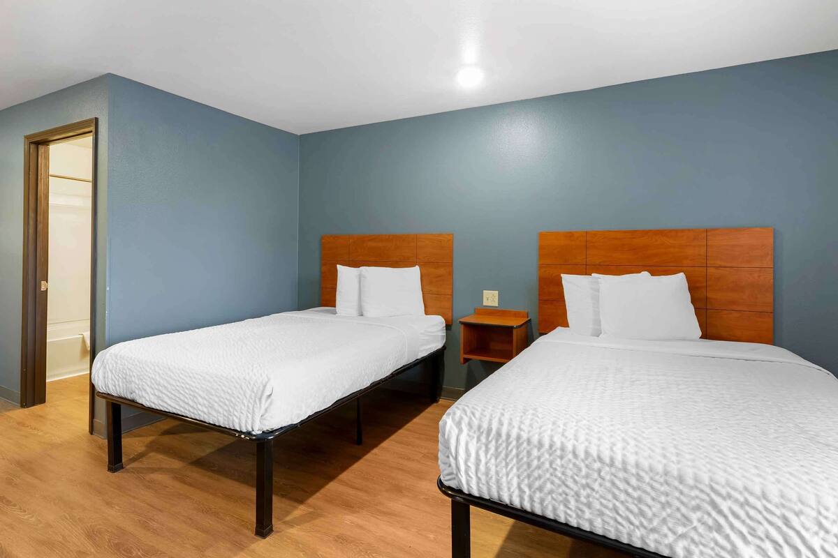 A hotel room with two single beds, wooden headboards, white bedding, a small wooden nightstand, and a door to the left.