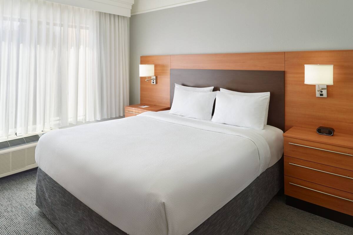 Une chambre d’hôtel avec un grand lit, deux tables de chevet, une petite table et une fenêtre avec des rideaux légers.