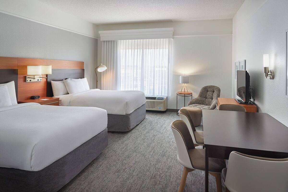 Une chambre d’hôtel avec deux lits, un bureau, une chaise, une télévision et une fenêtre avec des rideaux.