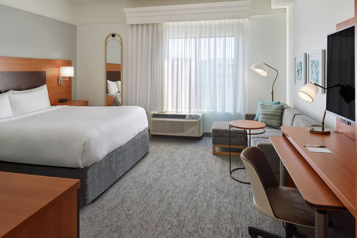 Une chambre d’hôtel avec un grand lit, un bureau, une chaise, une petite table, un appareil de climatisation et une grande fenêtre avec des rideaux légers.