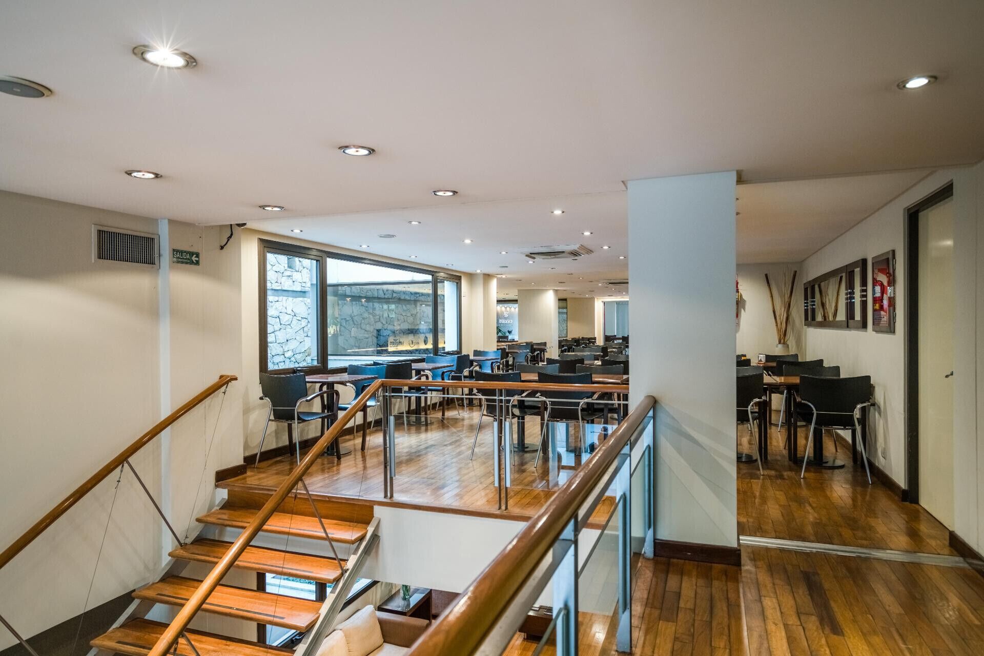Un interior moderno con una escalera, piso de madera y una zona de estar con mesas y sillas.