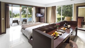 A modern hotel room with a large bed, a desk with fruit, a TV, and a balcony with a view of the outdoors.