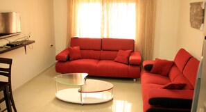 A living room with a red sofa, a glass coffee table, and a TV on a stand. - Uptown Modern Furnished Apartments (Amman)
