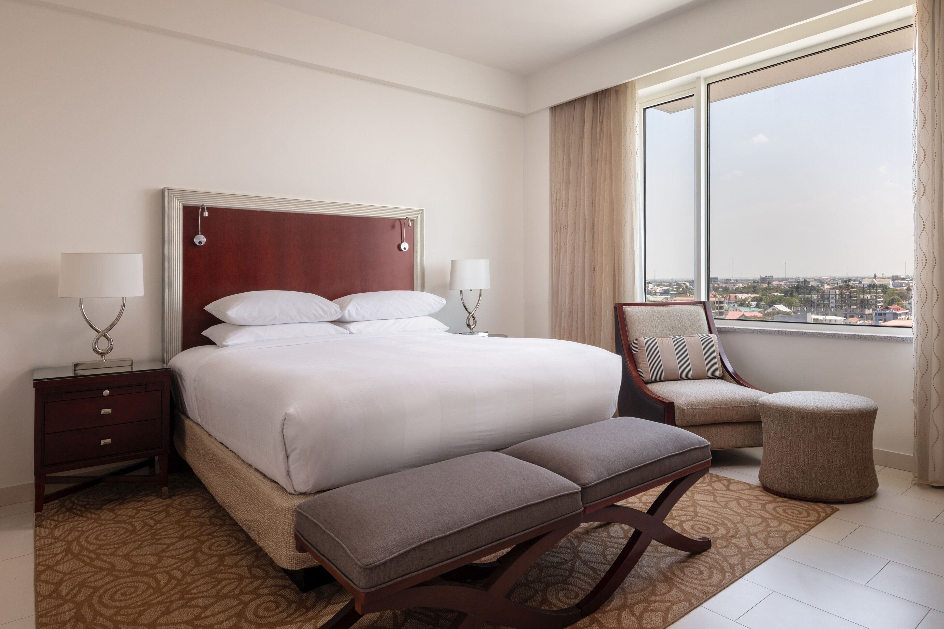 A hotel room with a large bed, a bedside table with lamps, a chair, a small ottoman, and a view of the city through the window.