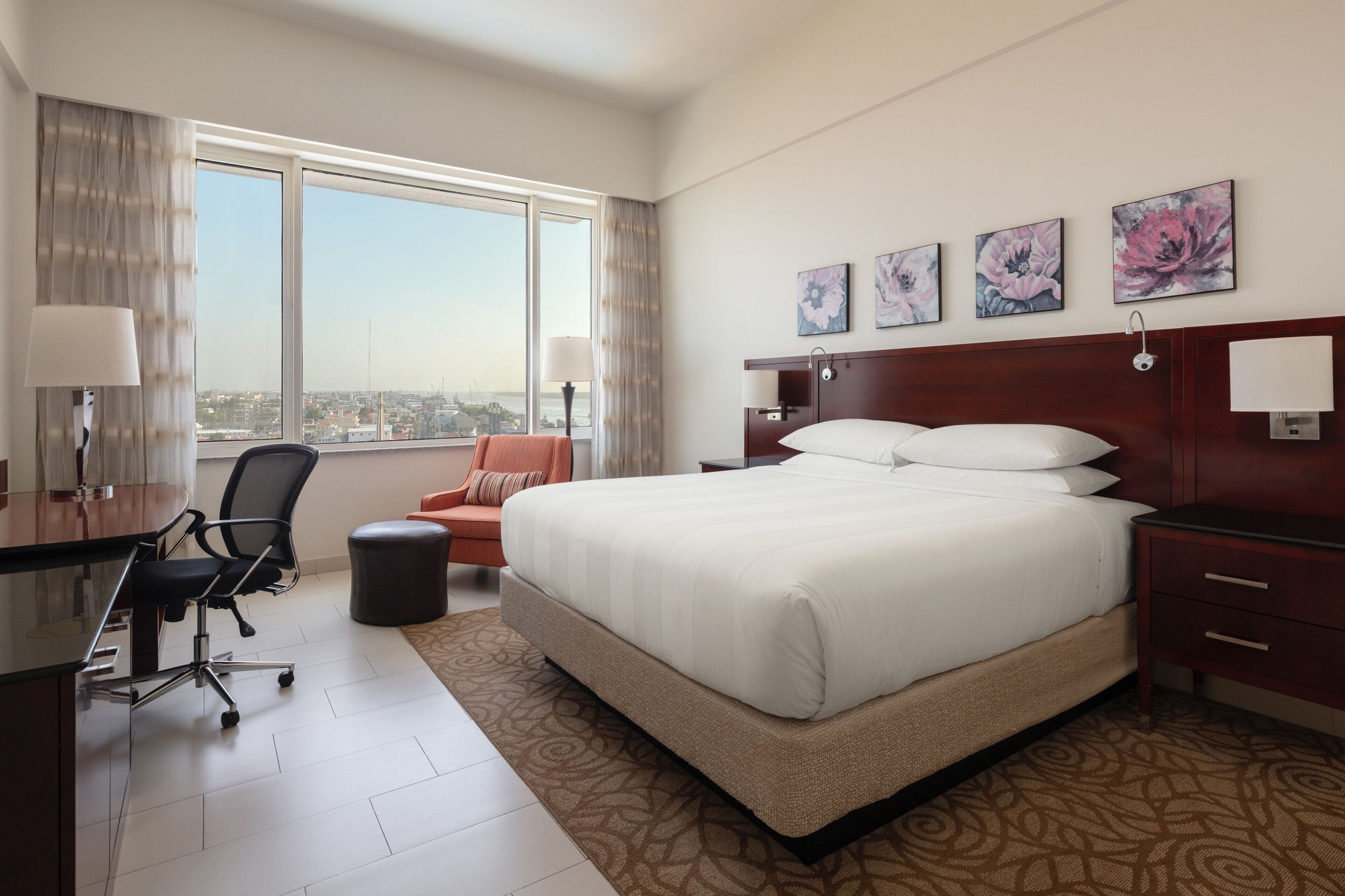 A hotel room with a large bed, a desk with a chair, a view of the city through large windows, and floral artwork on the wall.