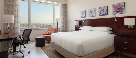 A hotel room with a large bed, a desk with a chair, a view of the city through large windows, and floral artwork on the wall.
