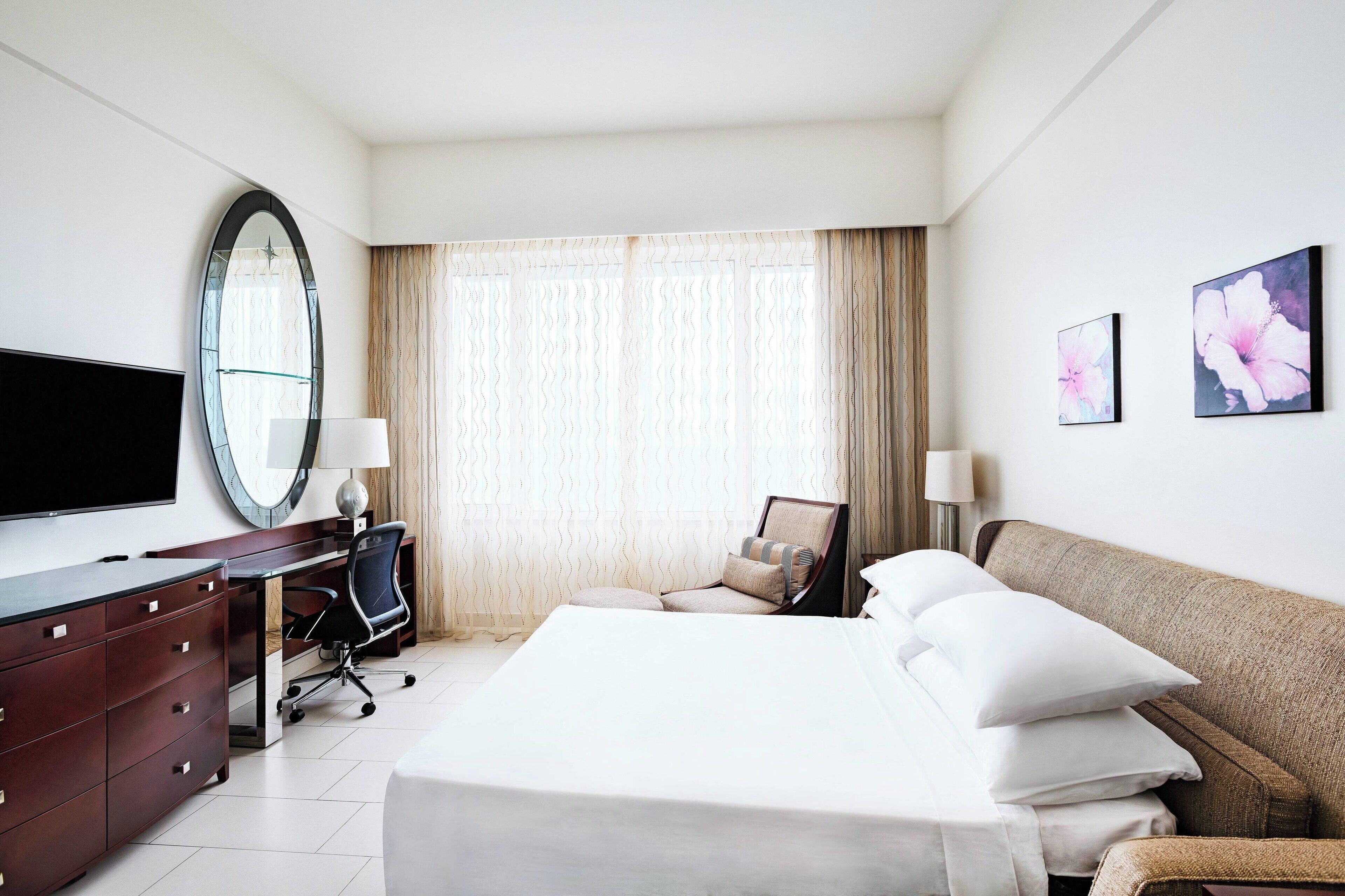 Une chambre d’hôtel avec un lit, un bureau, une chaise, une télévision, un miroir et deux tableaux accrochés au mur.