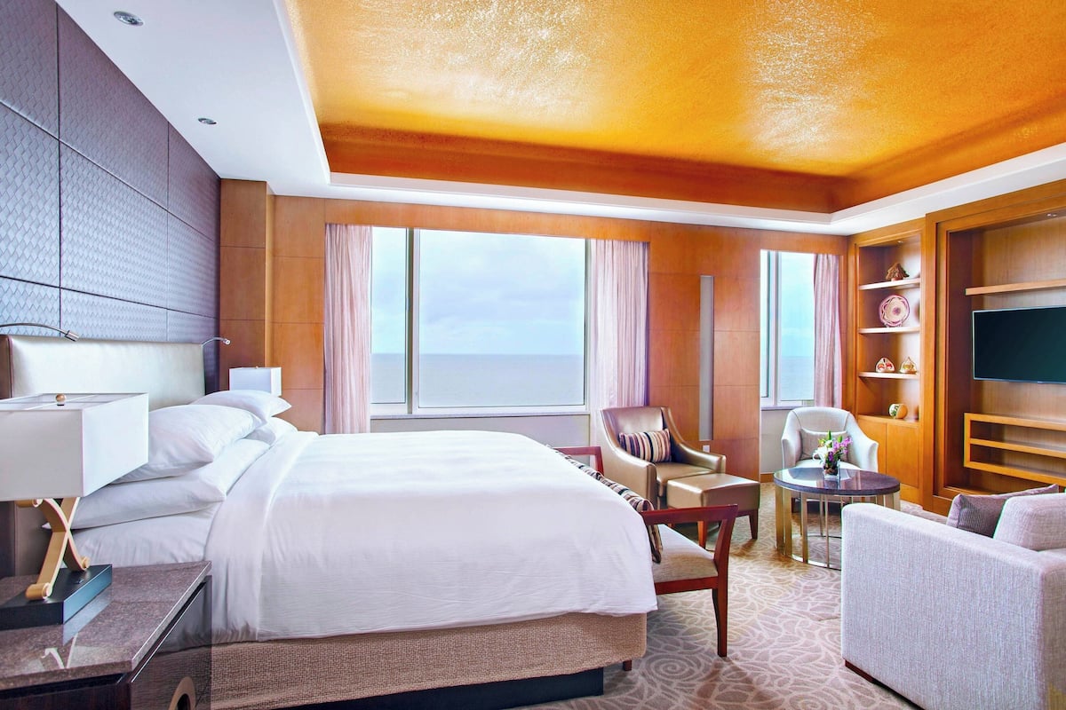 A hotel room with a large bed, two armchairs, a small table, a TV, and a large window with curtains.