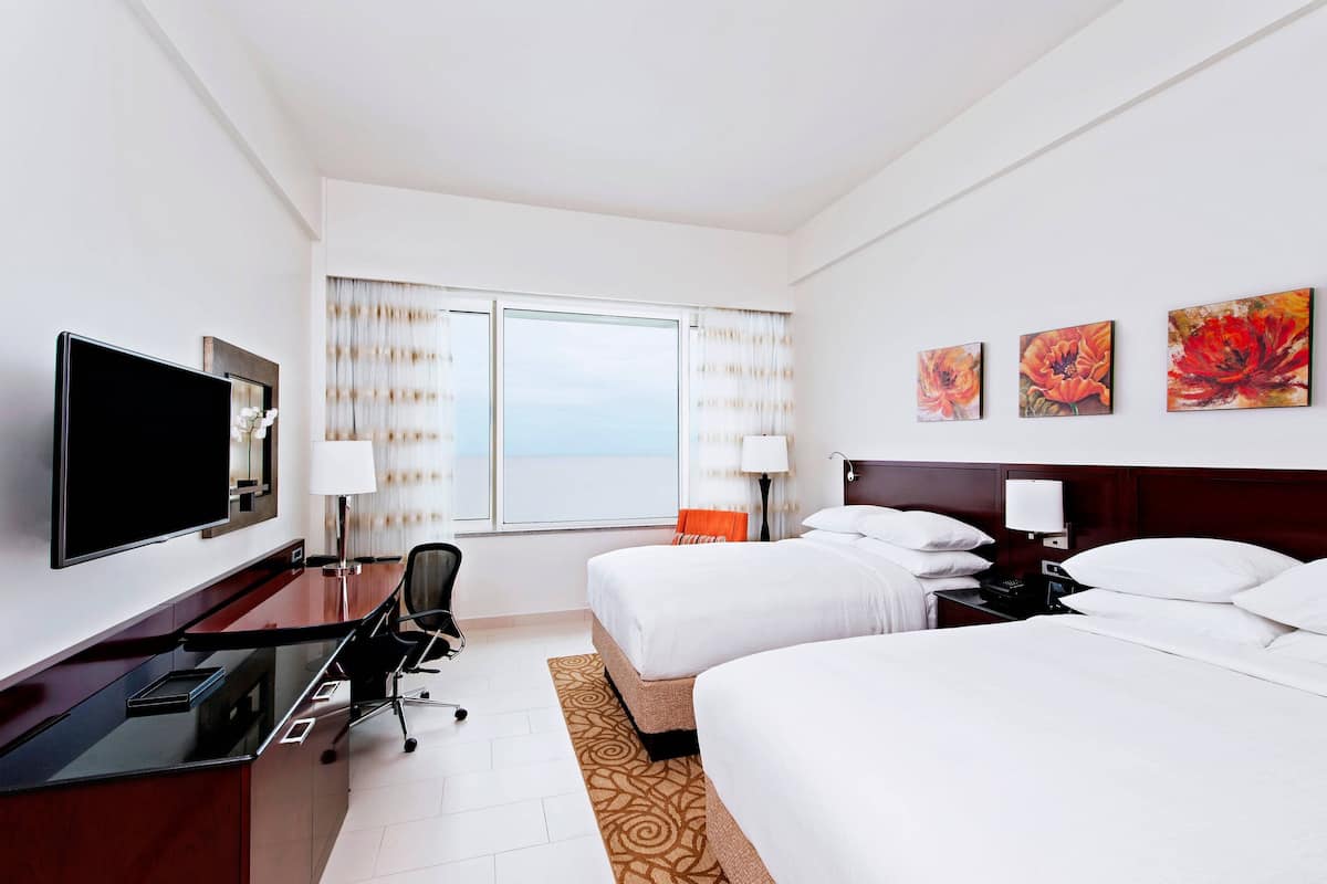 A hotel room with a bed, a desk with a TV, a chair, a nightstand, and a window with curtains.