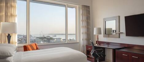 A hotel room with two beds, a desk, a chair, a TV, and a large window with a view of the city and water.