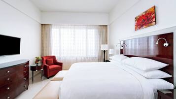 A hotel room with a large bed, a TV, a red armchair, a small table with a vase, and a painting on the wall.