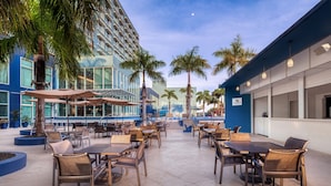 An outdoor patio area with tables and chairs, palm trees, and a modern building in the background.