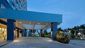 A modern hotel entrance with a covered walkway, palm trees, and a well-lit facade.