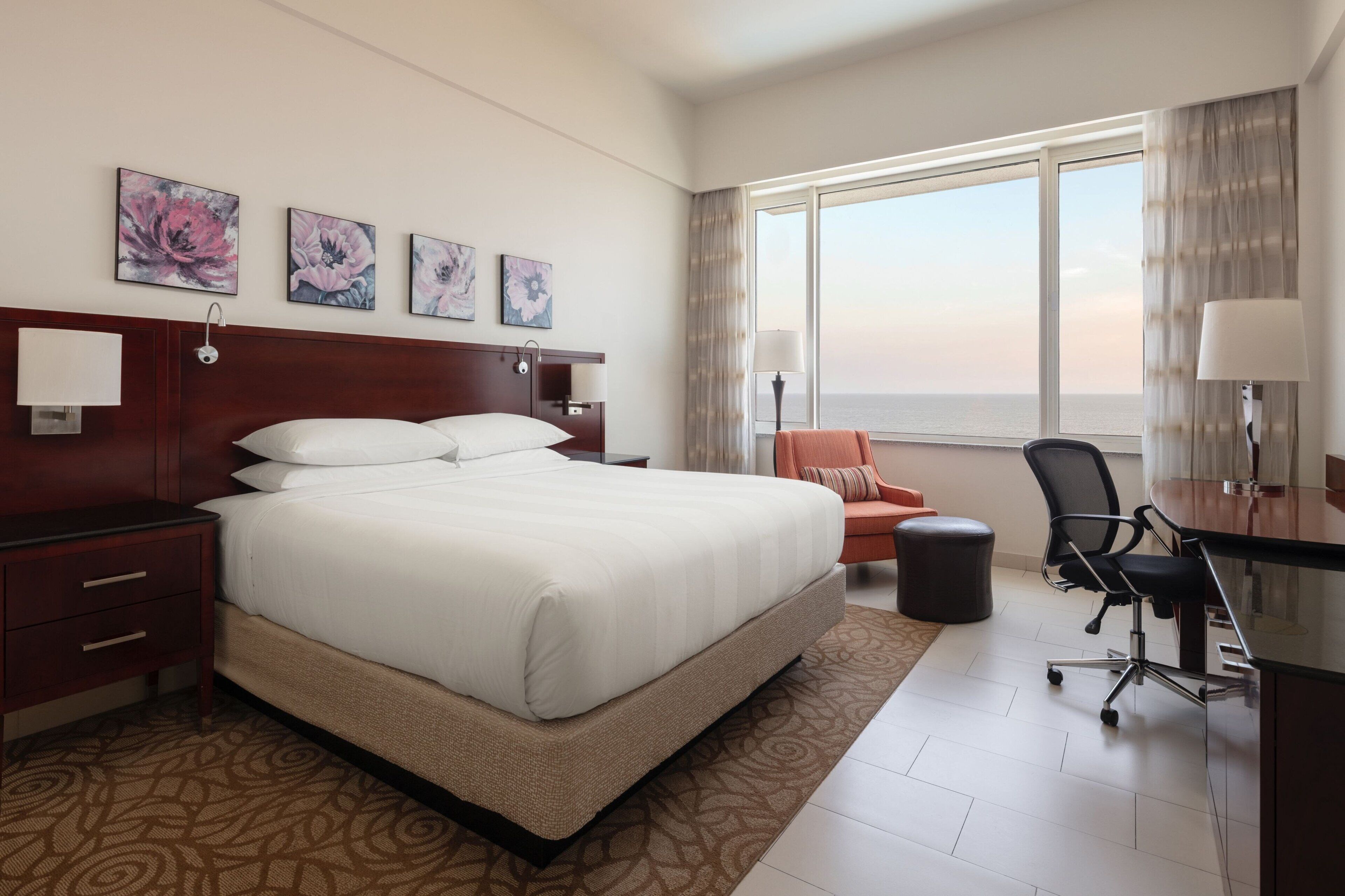 Une chambre d’hôtel avec un grand lit, un bureau avec une chaise, une vue sur la mer et des œuvres d’art accrochées au mur.