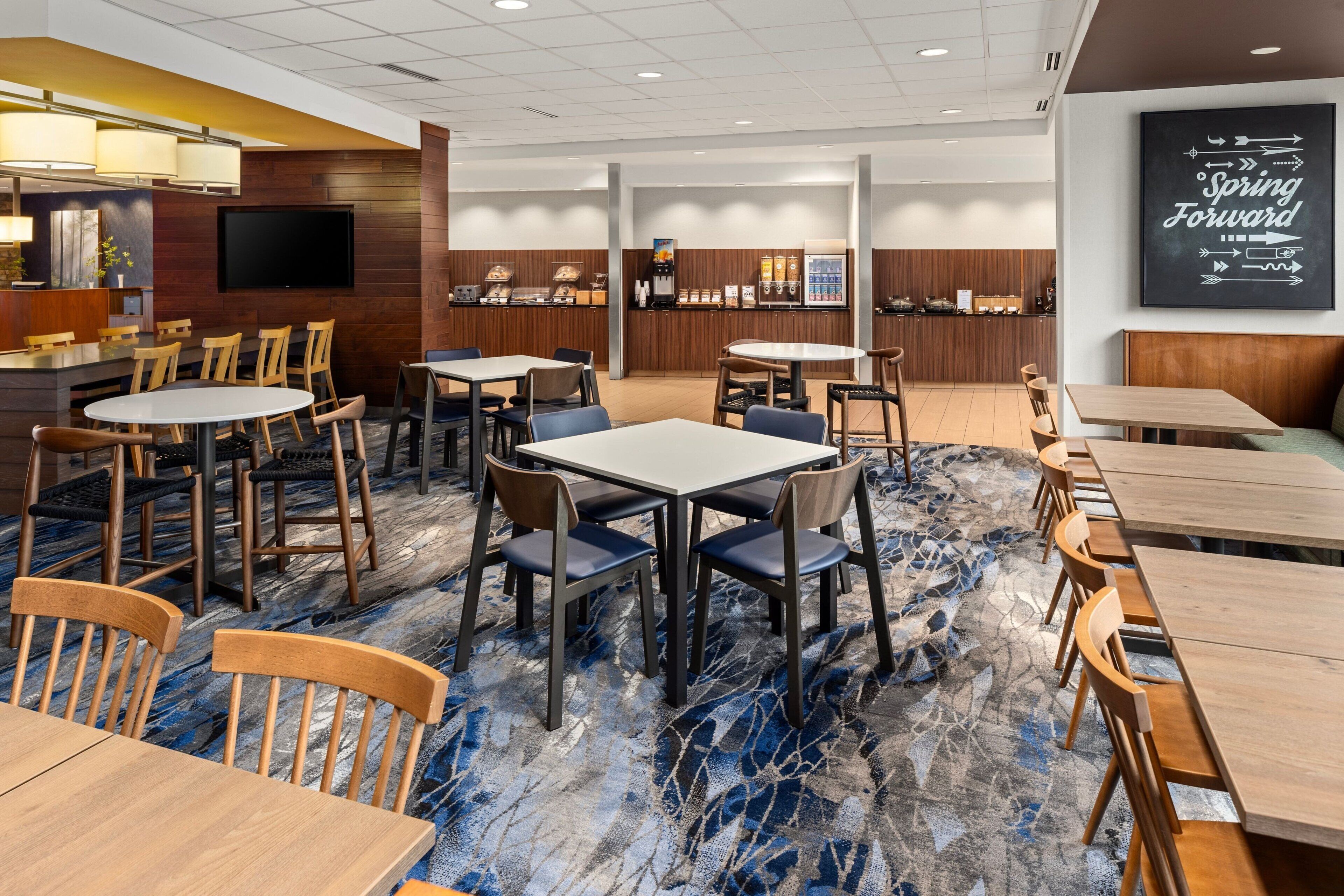 A hotel dining area with multiple tables and chairs, a television mounted on the wall, and a 'Spring Forward' sign.