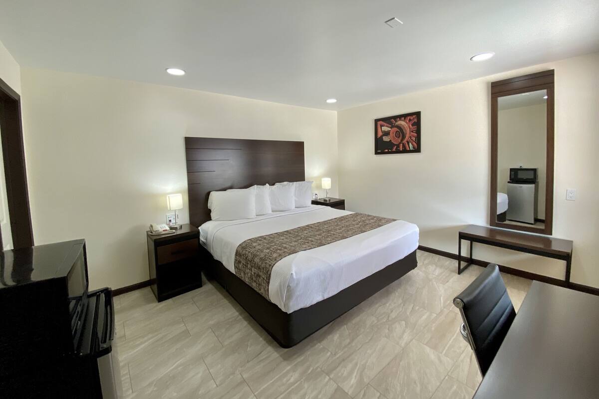 A hotel room with a large bed, bedside tables, a desk, a chair, a mirror, and a wall-mounted artwork.
