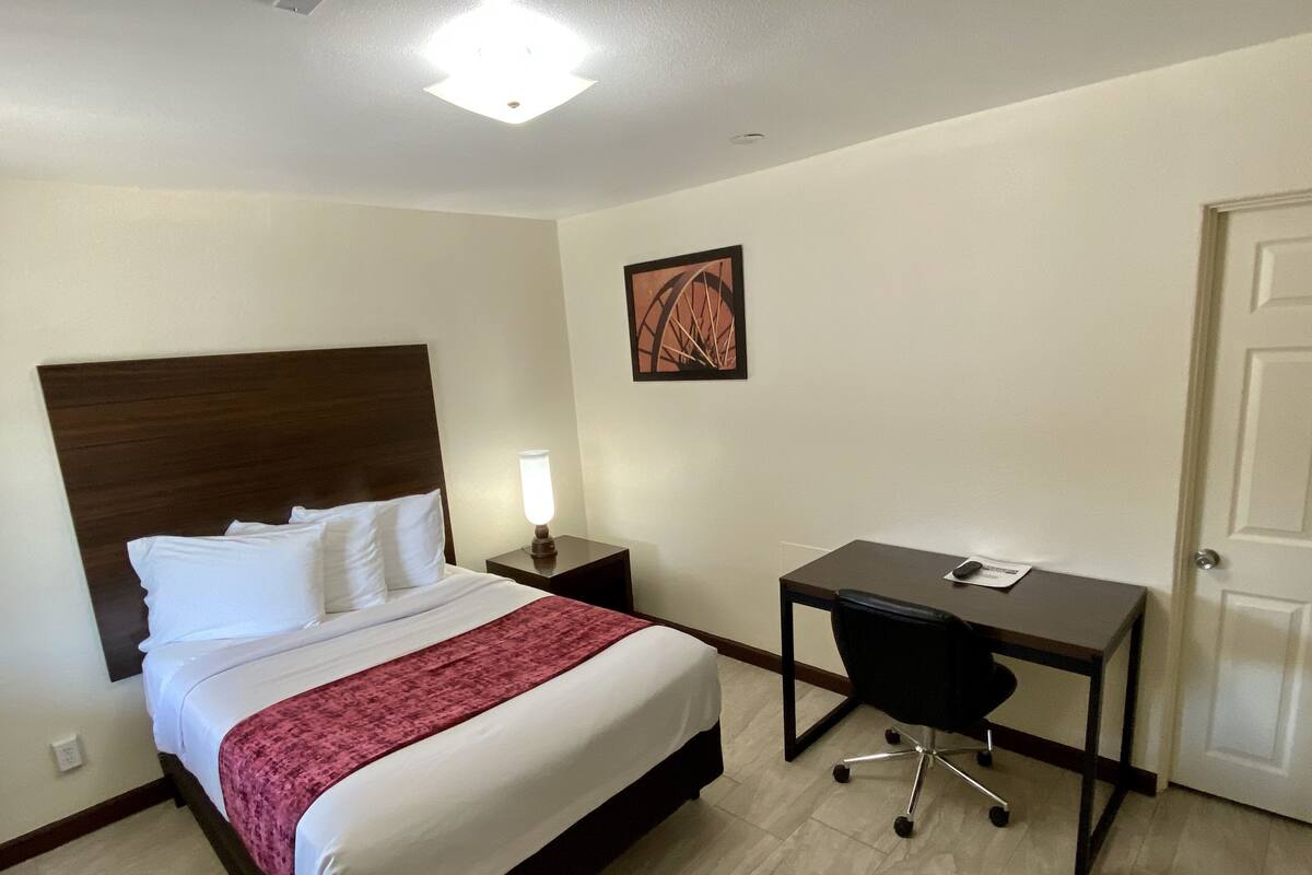 A hotel room with a bed, a desk, a chair, a nightstand, a lamp, and a door.
